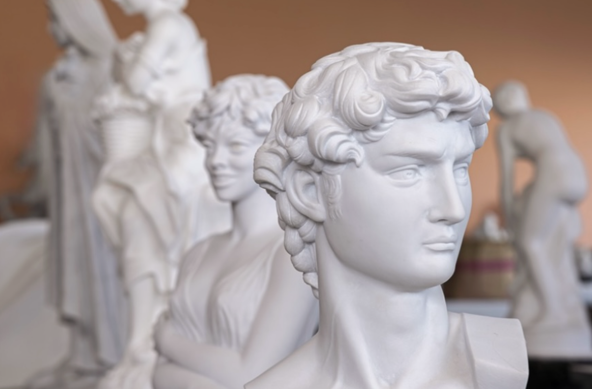 Close-up of a classical marble bust of a man with curly hair, showcasing timeless beauty. In the background, other sculptures, including a female figure, are blurred. The setting appears to be an art studio or gallery with a warm-toned wall.